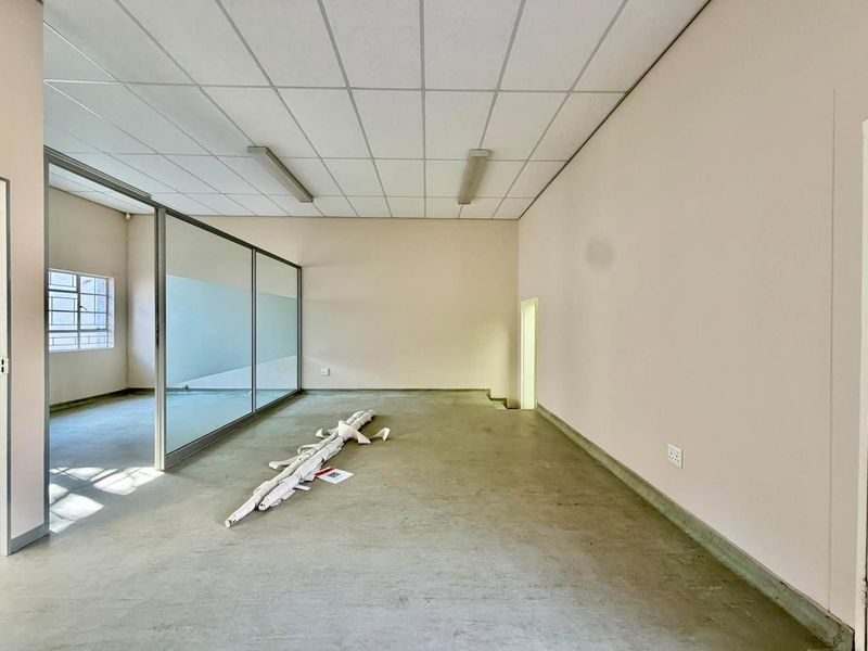 An empty, open-plan office space featuring a glass partition, suspended ceiling with fluorescent lighting, and concrete flooring.