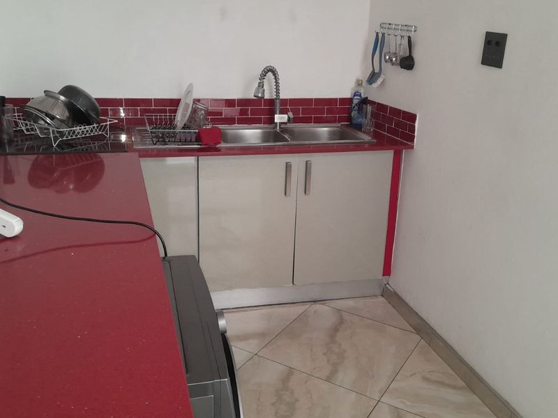 A compact kitchen area featuring red countertops, a matching tiled backsplash, double basin sink with gooseneck faucet, and neutral cabinetry.