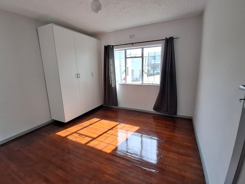 A bright, empty bedroom featuring polished parquet flooring, a large built-in wardrobe, and a metal-framed window allowing for natural light.