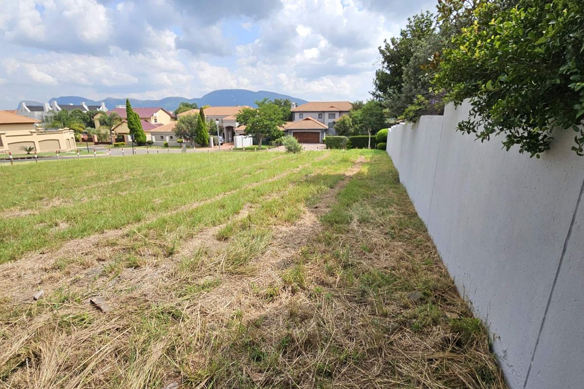 A vacant residential plot featuring a boundary wall, grassy terrain, and views of the surrounding neighborhood and distant mountains.