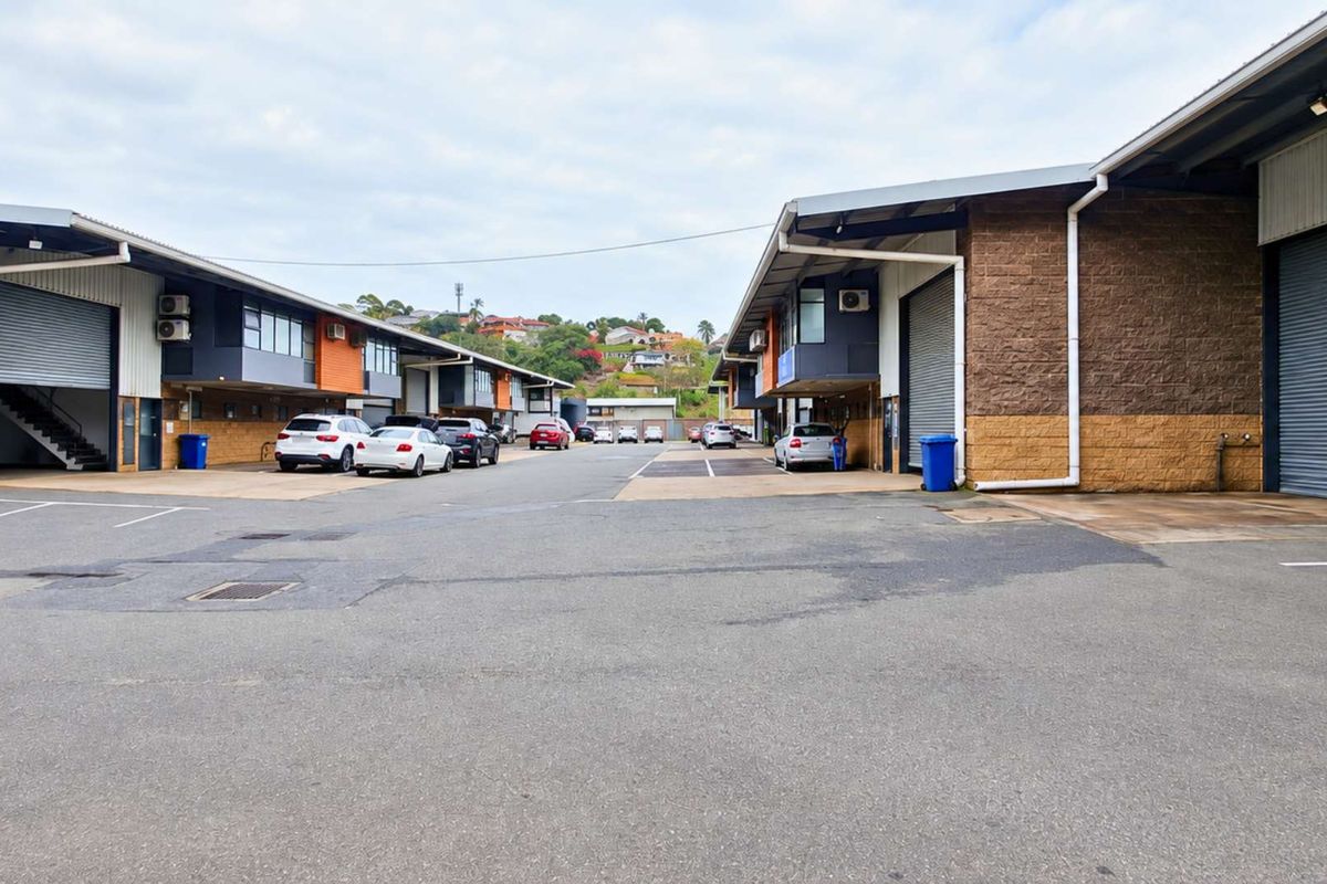 Exterior view of an industrial complex featuring multiple units with roller shutter doors, brick facades, and a shared asphalt driveway.