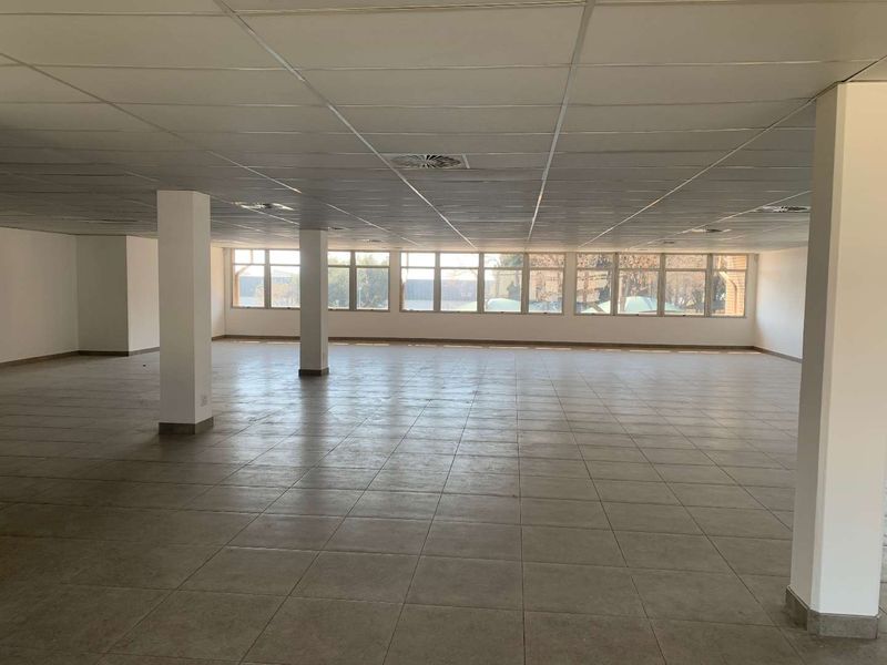 Spacious open-plan industrial office area featuring large windows, neutral ceramic tile flooring, and a suspended ceiling with integrated structural columns.