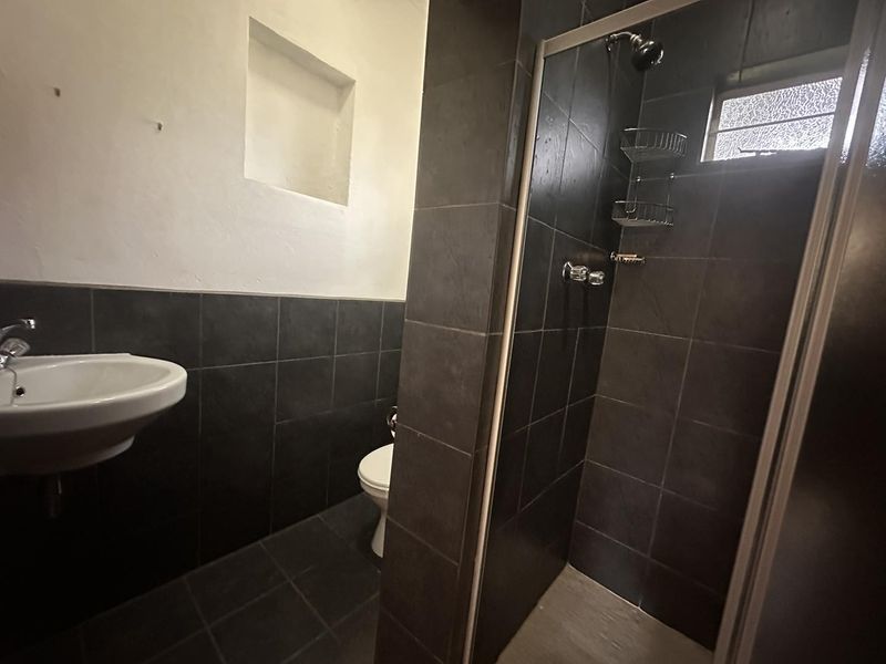 A functional bathroom featuring a shower enclosure, wall-mounted basin, and toilet with dark tiled finishes and a small obscured glass window.