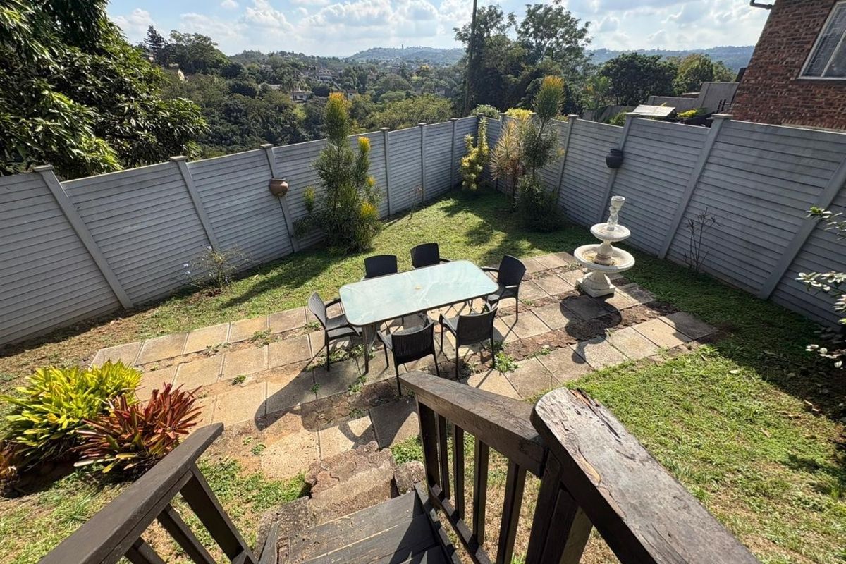 A rear garden featuring a paved patio area, a timber staircase, and a stone fountain, enclosed by a precast concrete boundary wall.