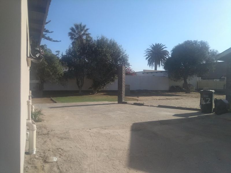 A rear exterior view of the property featuring a concrete paved area, boundary walls, and an open garden space with mature trees.