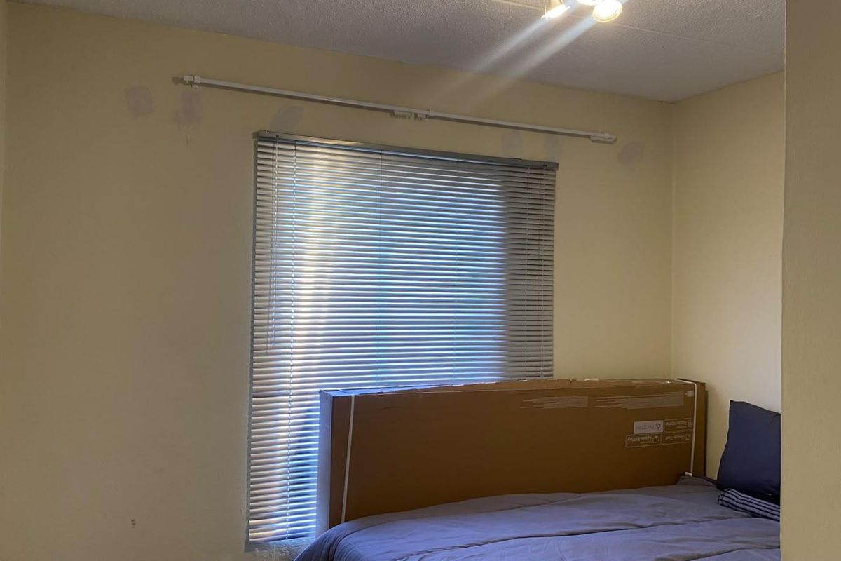 A bedroom featuring a window with venetian blinds and a curtain rod, showing visible wall surface imperfections and patchy paintwork.
