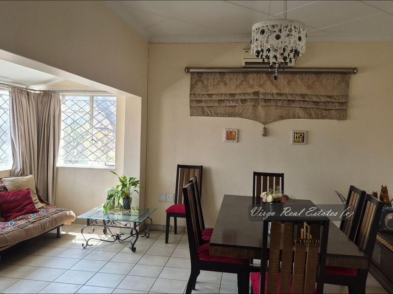 A dining area featuring tiled flooring, a wall-mounted air conditioning unit, and large windows with security bars, providing a functional space for meals.