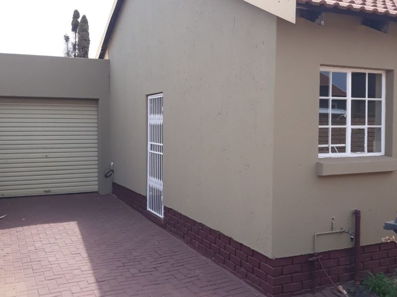 Exterior view of a townhouse featuring a paved driveway, roller garage door, security gate, and a tiled roof with brick plinth detailing.
