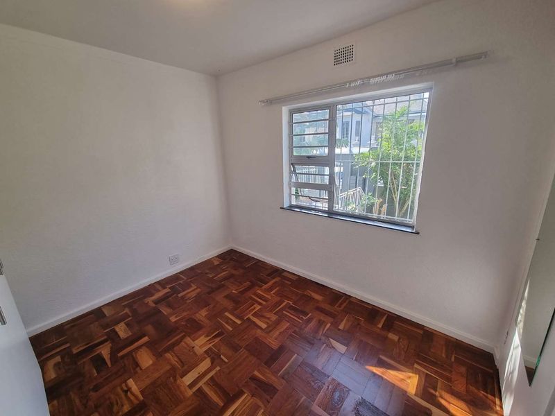An empty bedroom featuring classic parquet flooring, a louvered window with security bars, and a neutral white wall finish.