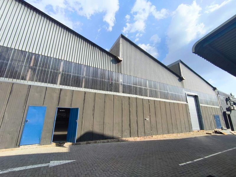 Exterior view of an industrial warehouse featuring corrugated metal cladding, translucent wall panels, a large roller shutter door, and a paved driveway.