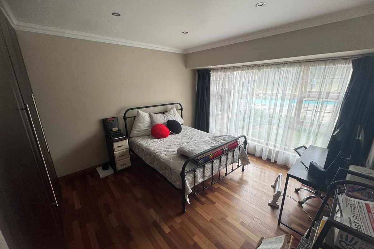 A well-lit bedroom featuring warm-toned wooden flooring, built-in cupboards, and large windows providing natural light and a view of the outdoor pool area.