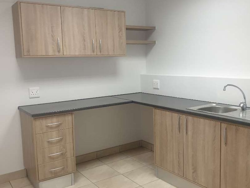 A clean and functional scullery or utility area featuring light wood-finish cabinetry, dark countertops, a stainless steel sink, and neutral floor tiling.