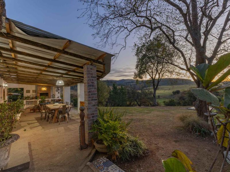 A spacious covered patio featuring brick pillars, exposed timber rafters, and a built-in braai, overlooking a scenic garden and rural landscape.