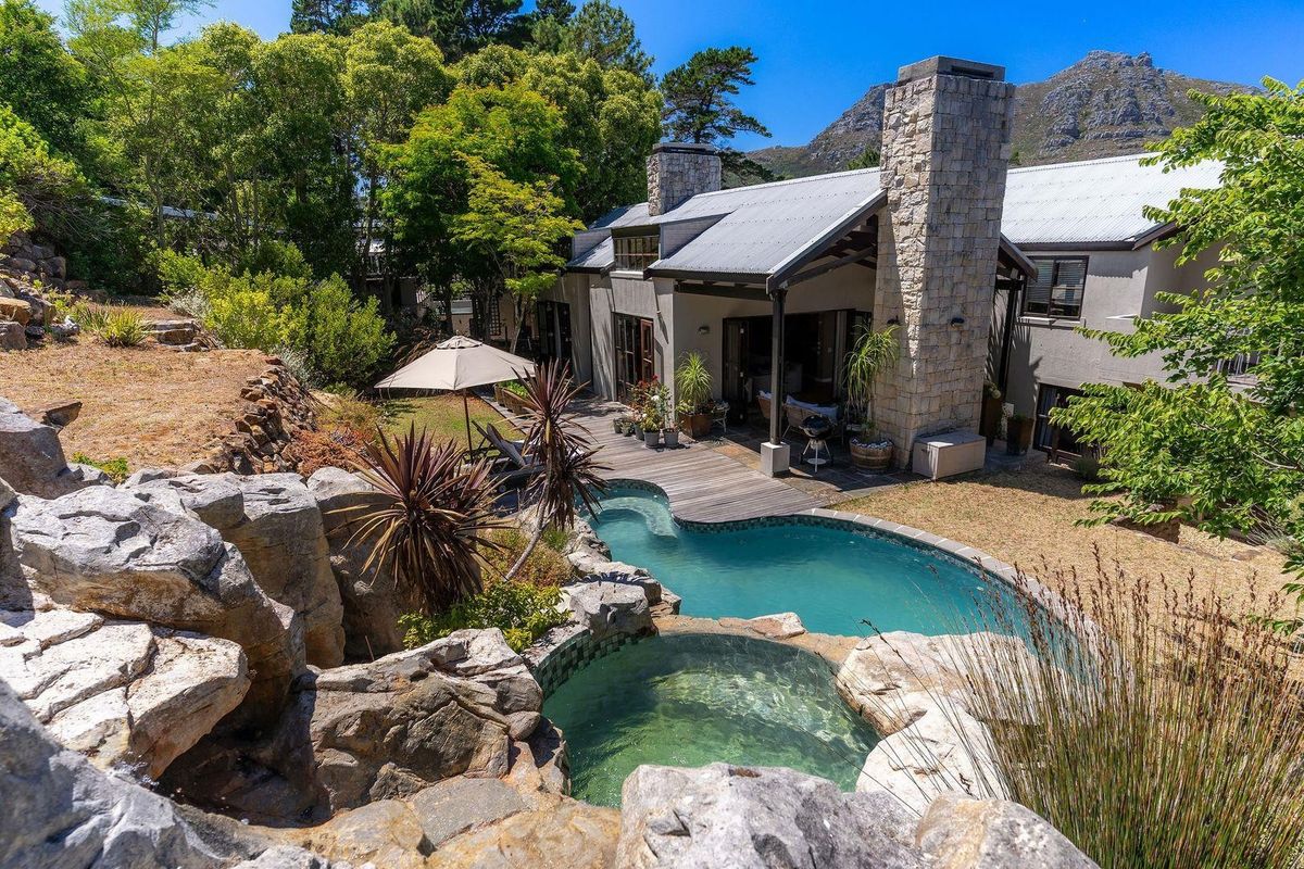 A scenic rear exterior view featuring a natural rock-integrated swimming pool, wooden decking, and a stone chimney set against a mountainous backdrop.