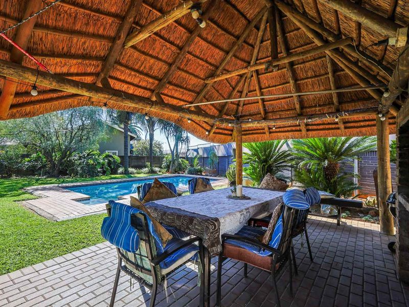 Covered outdoor patio featuring a traditional thatched roof, brick paving, and a built-in braai area overlooking the swimming pool and garden.