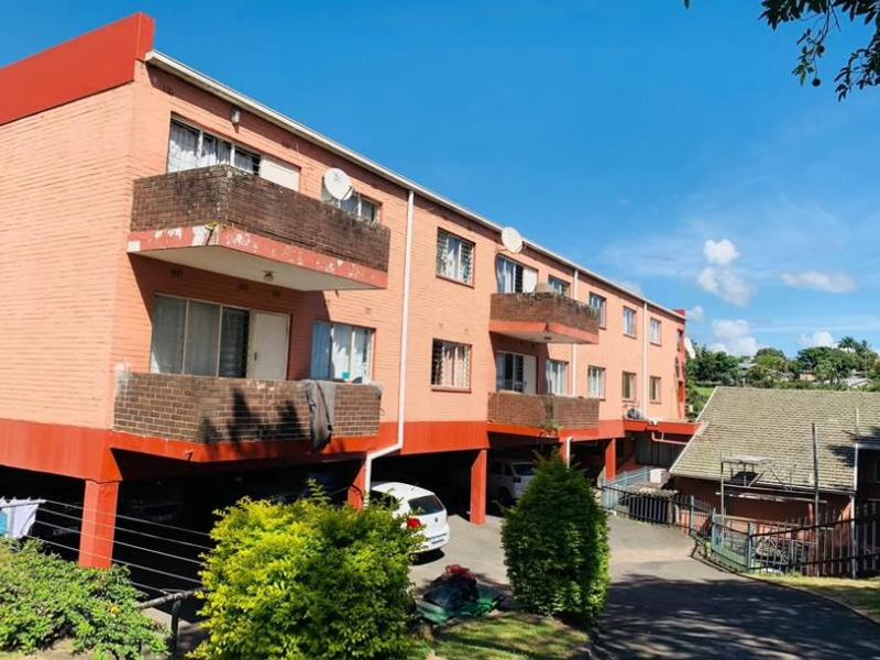 Rear view of a multi-story apartment building featuring brick facade, private balconies, and a ground-level covered parking area.