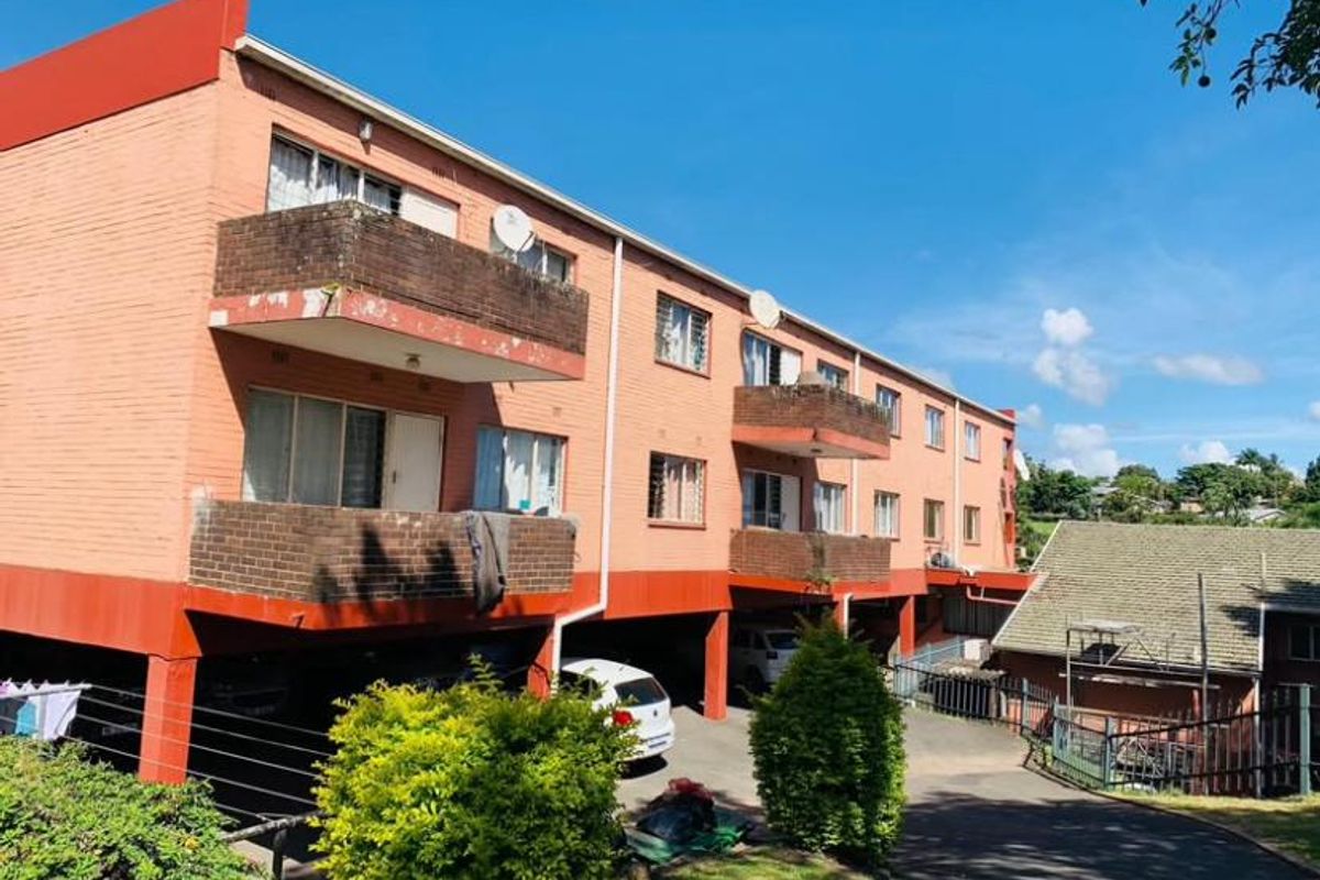 Rear view of a multi-story apartment building featuring brick facade, private balconies, and a ground-level covered parking area.