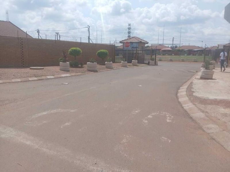 A street view showing the entrance to a residential estate with a brick boundary wall, security gate, and paved access road.