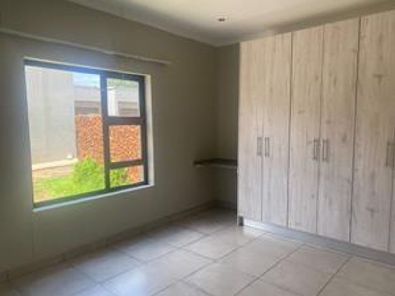 A bright, empty bedroom featuring neutral tiled flooring, modern built-in cupboards, and a large window providing natural light.
