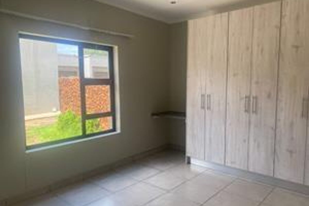 A bright, empty bedroom featuring neutral tiled flooring, modern built-in cupboards, and a large window providing natural light.
