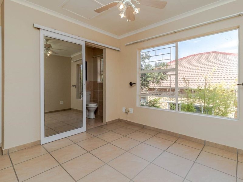 A bright, empty bedroom featuring tiled flooring, a ceiling fan, and an en-suite bathroom with a sliding mirror door.