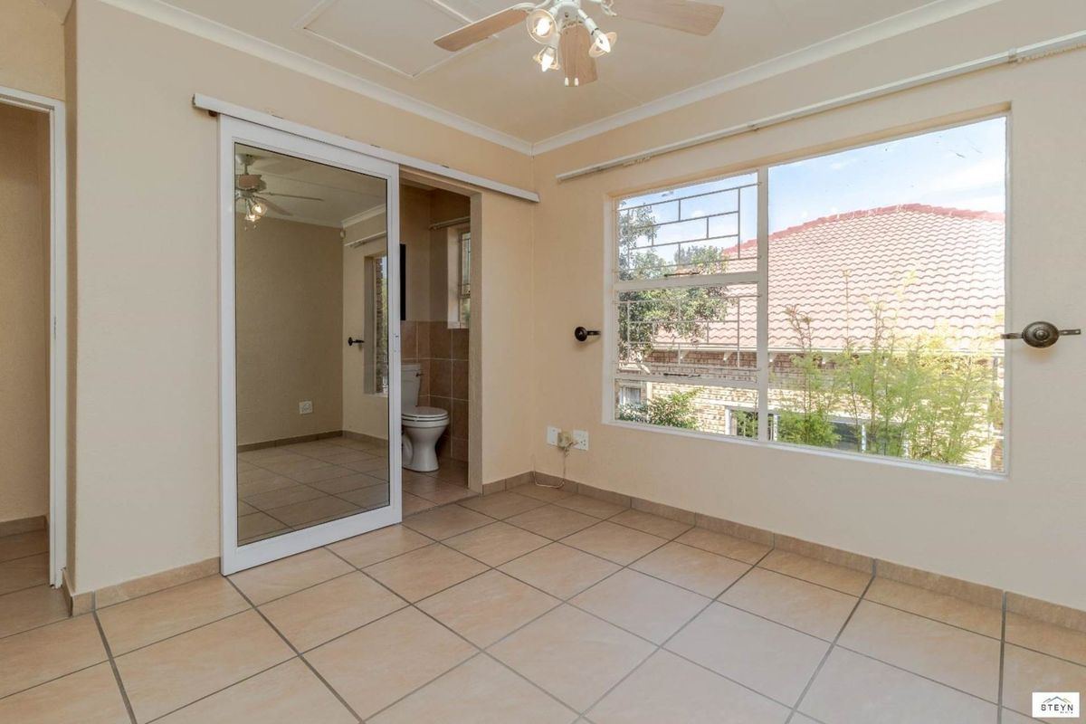 A bright, empty bedroom featuring tiled flooring, a ceiling fan, and an en-suite bathroom with a sliding mirror door.