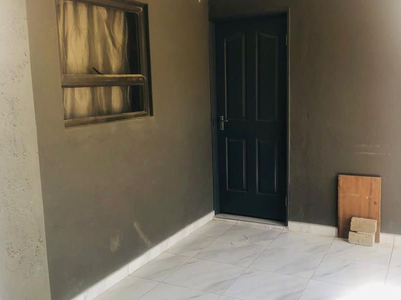 A covered patio area featuring tiled flooring, a dark-painted exterior door, and a window, presenting a clean but basic outdoor space.