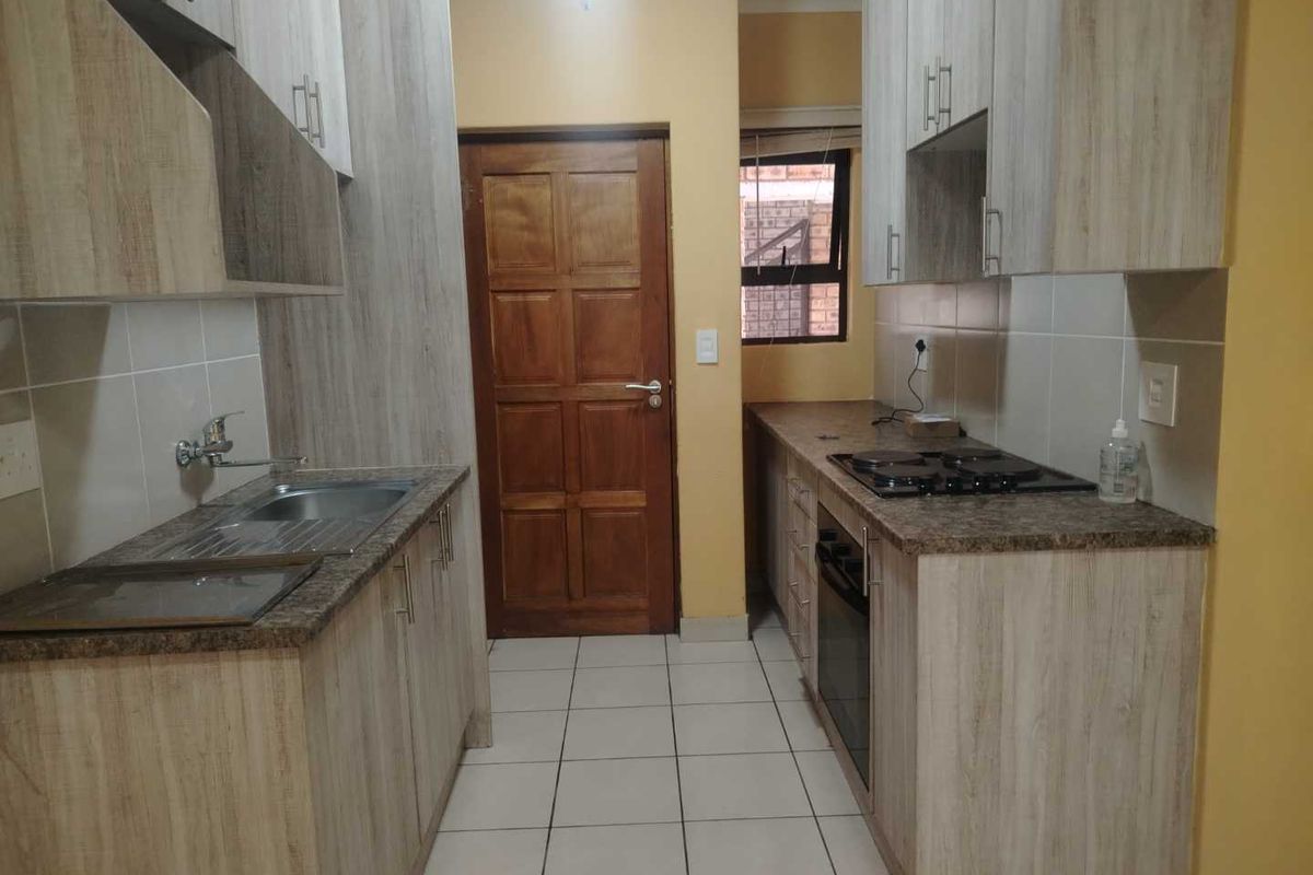 A functional galley-style kitchen featuring light wood-grain cabinetry, granite countertops, built-in oven and hob, and tiled flooring.