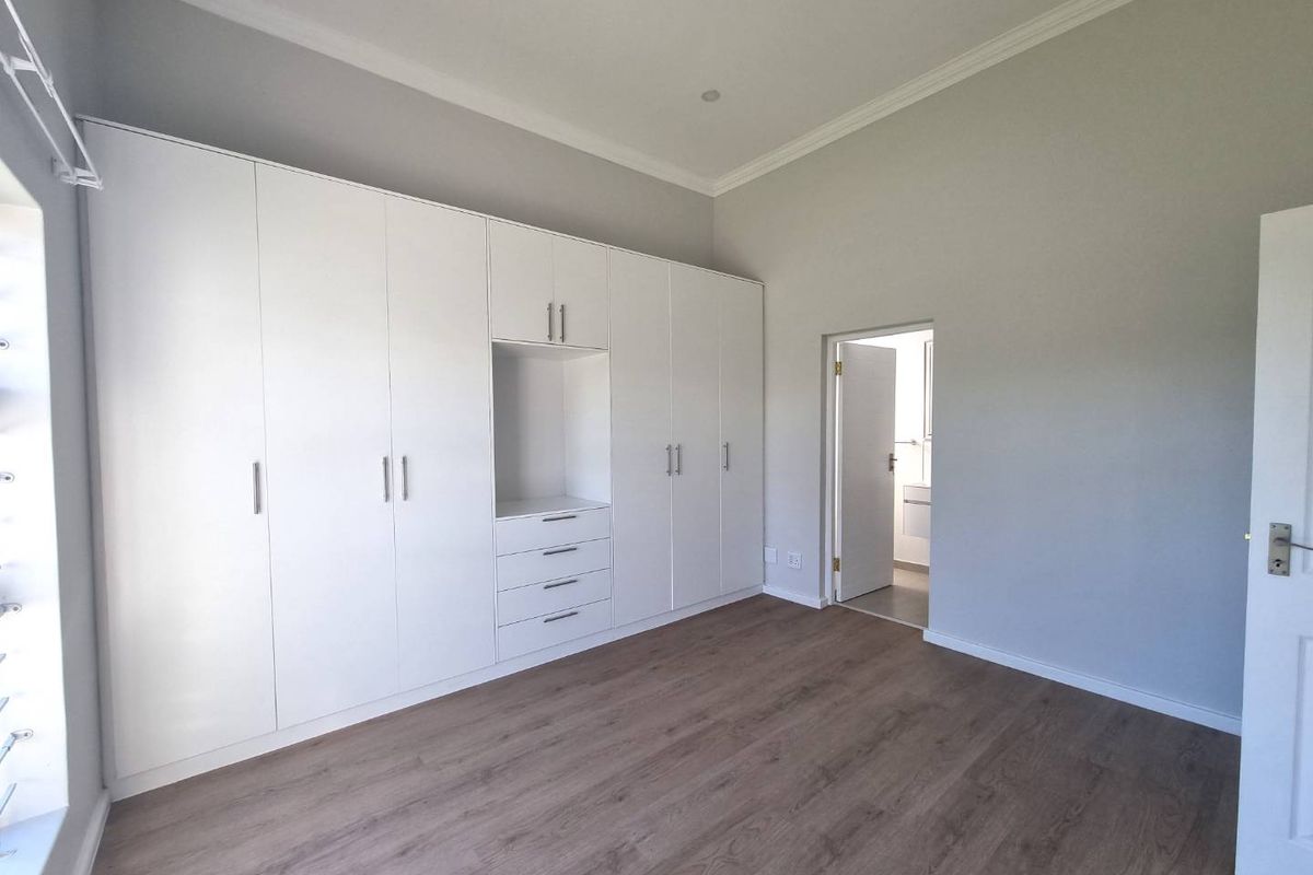 A bright, empty bedroom featuring modern white built-in cupboards, laminate flooring, and neutral wall finishes with access to an adjoining room.