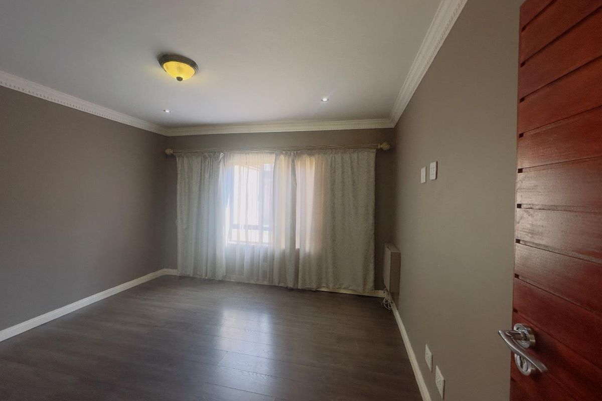 A spacious, empty bedroom featuring dark laminate flooring, neutral wall tones, decorative crown molding, and a large window with installed curtain rods.