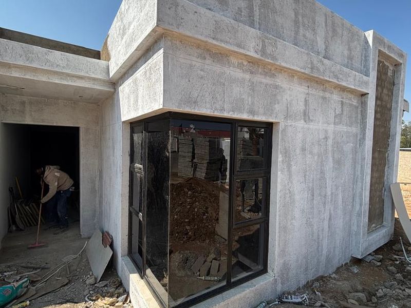 Exterior view of a house under construction featuring a modern corner window design and unfinished cement plaster walls.