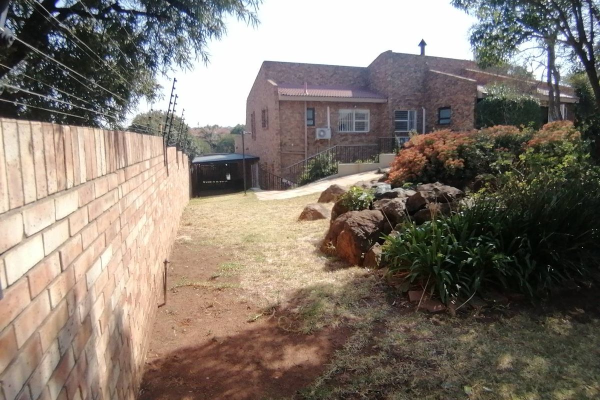 Rear view of a face brick house featuring a garden area, boundary wall with electric fencing, and a paved walkway leading to a carport.