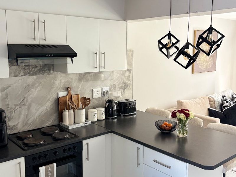 Modern open-plan kitchen featuring white cabinetry, dark countertops, integrated appliances, and a breakfast bar, seamlessly flowing into the adjacent living area.