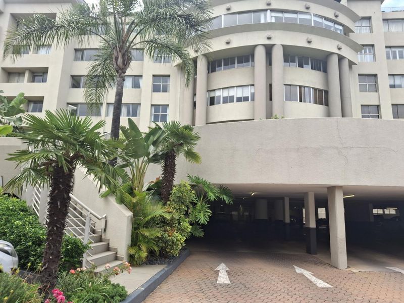 Front elevation of a multi-story apartment building featuring a covered parking entrance, paved driveway, and external staircase with landscaped surroundings.