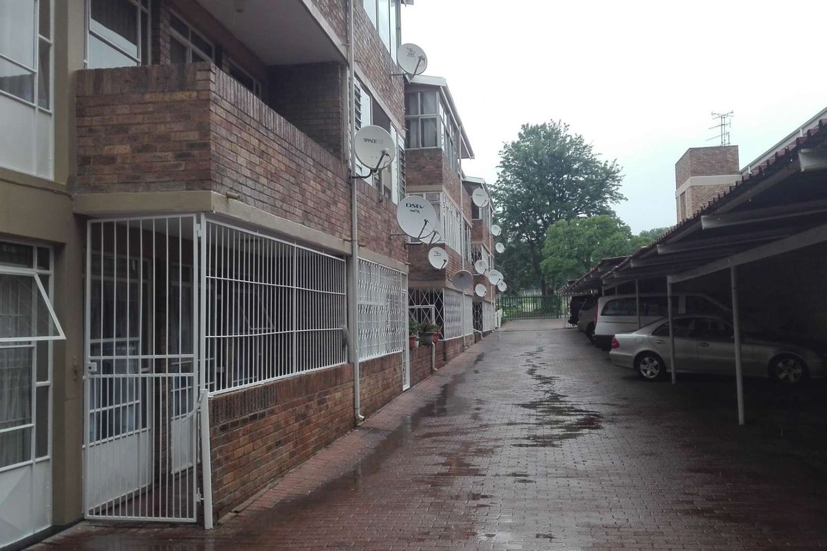 Exterior view of an apartment complex featuring brick facade, security gates, and covered parking bays along a paved driveway.