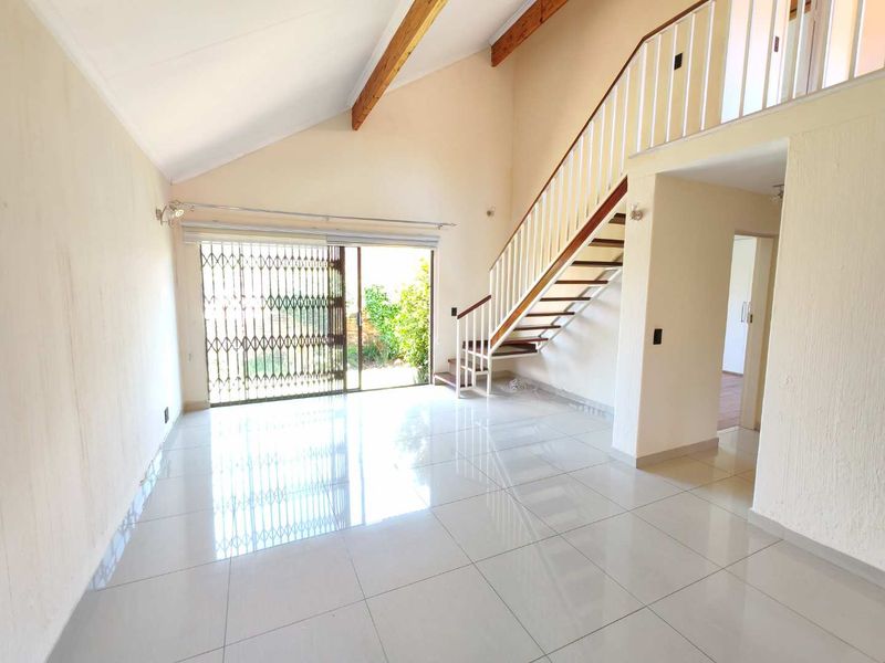 Spacious open-plan living area featuring high vaulted ceilings with exposed timber beams, tiled flooring, and a staircase leading to a mezzanine level.