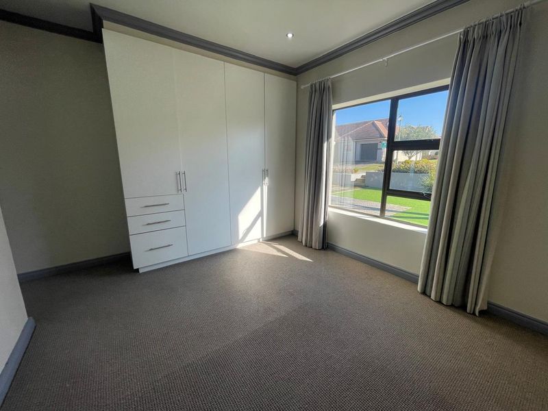A bright, empty bedroom featuring neutral carpeting, built-in cupboards, and large windows with views of the garden.