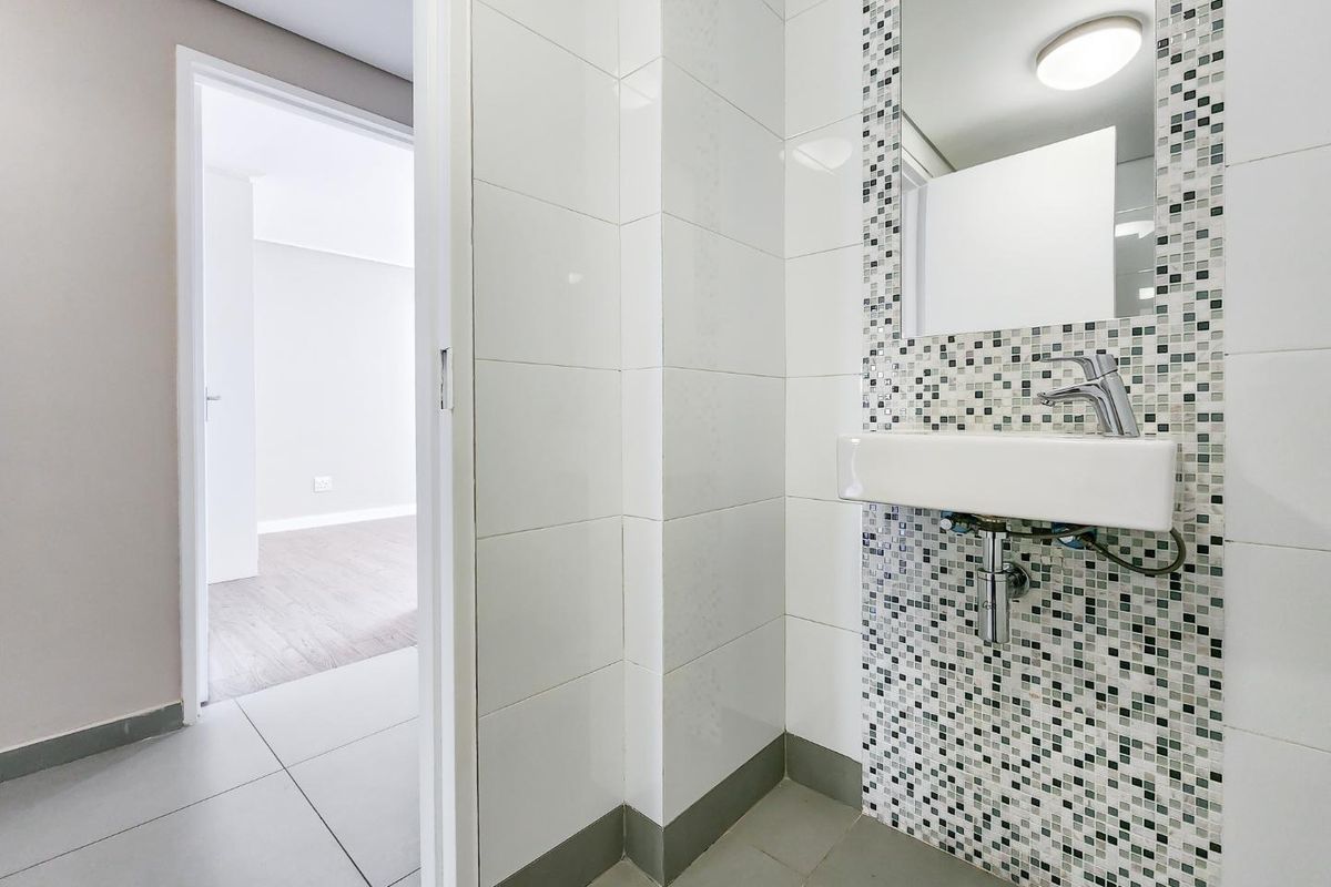 A clean, modern bathroom featuring a wall-mounted basin with a mosaic tile splashback and a built-in mirror, adjacent to an open living area.