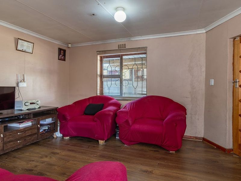 A living room featuring laminate flooring, a timber door, and a window with security bars, showing signs of wall staining and wear.