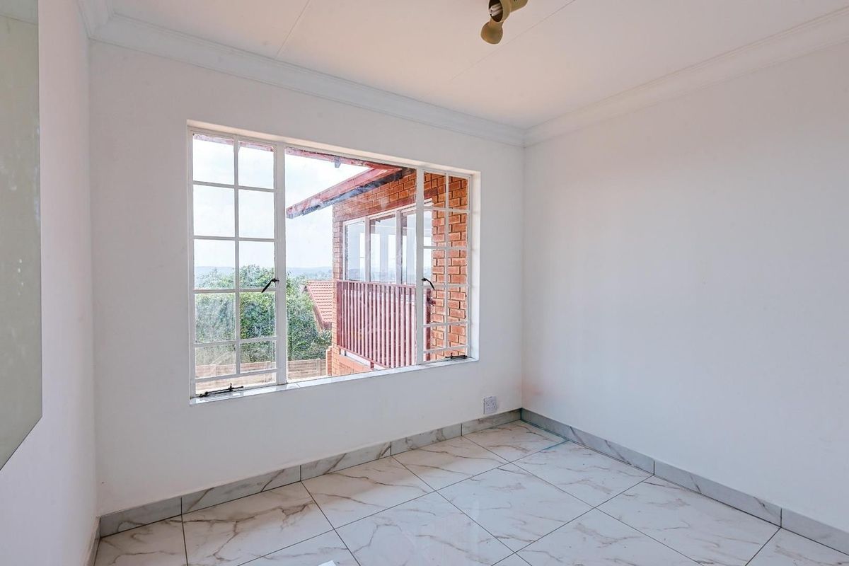 A bright, empty bedroom featuring white walls, marble-look tiled flooring, and a large window with views of the exterior brick facade.