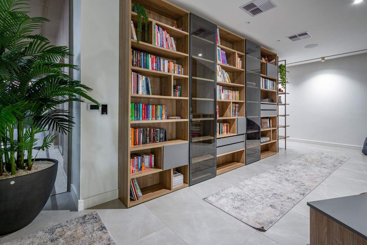 A modern living area featuring extensive floor-to-ceiling built-in timber shelving with integrated glass-fronted cabinets and recessed ceiling lighting.