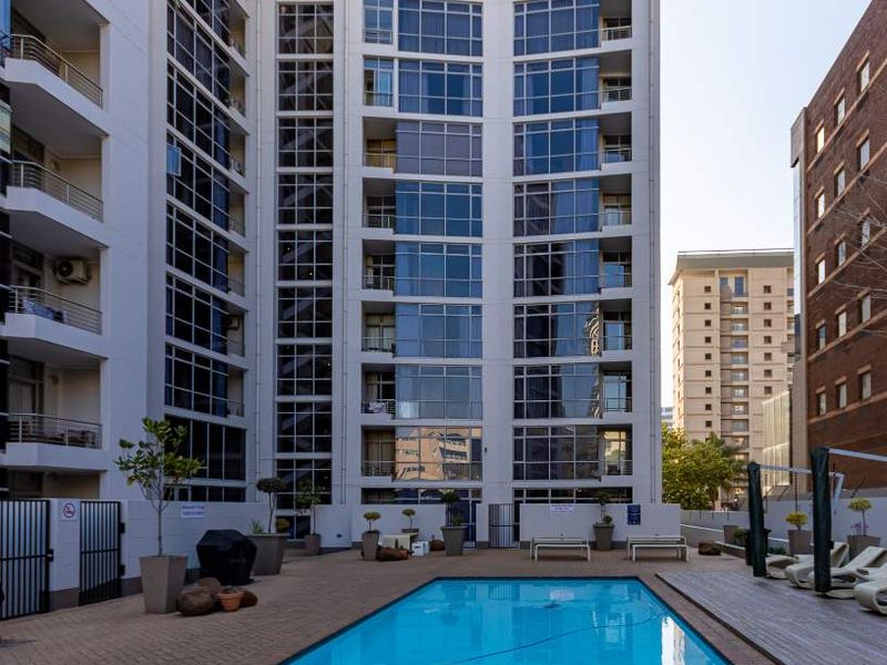 A well-maintained outdoor swimming pool area set within a modern high-rise apartment complex, featuring paved decking and glass-fronted building architecture.