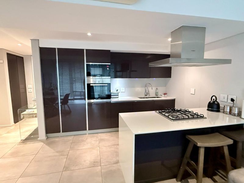 Modern kitchen featuring dark high-gloss cabinetry, stone countertops, a central island with a gas hob, and integrated stainless steel appliances.