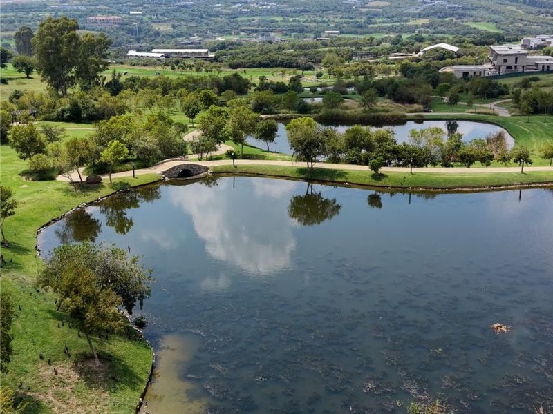 An elevated view of a landscaped residential estate featuring scenic ponds, walking paths, and a stone bridge set within a green parkland environment.