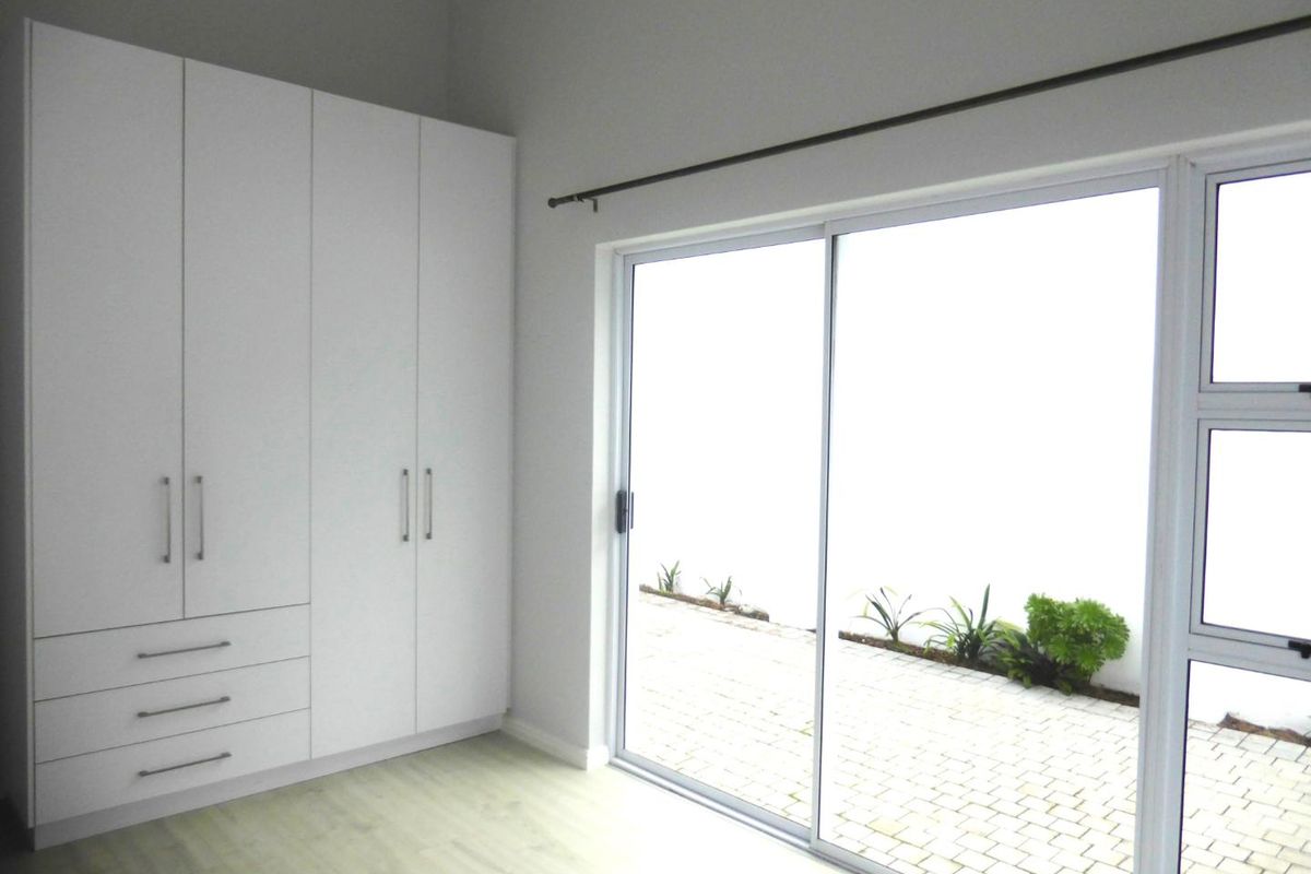 A bright, empty bedroom featuring modern white built-in cupboards, light laminate flooring, and a large sliding glass door providing access to the patio.