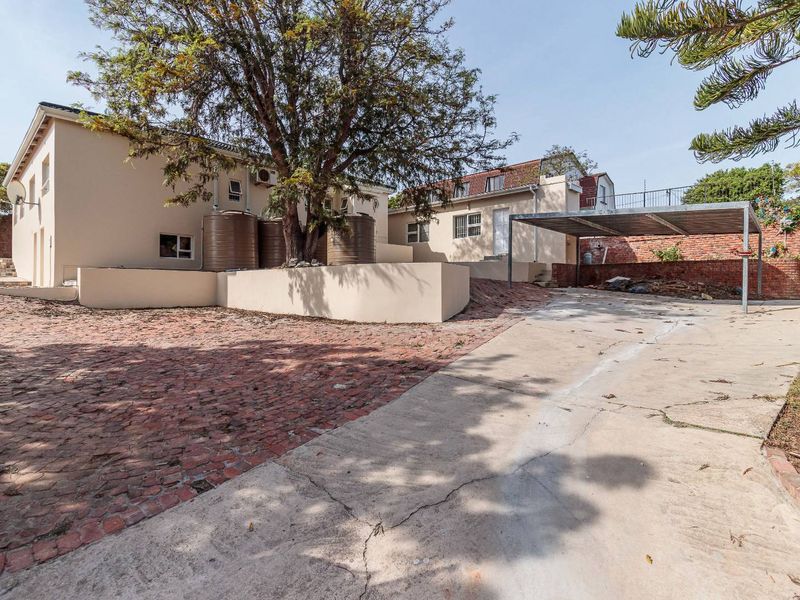 Rear exterior view featuring a large tree, water storage tanks, a carport, and a combination of brick and concrete driveway surfaces.