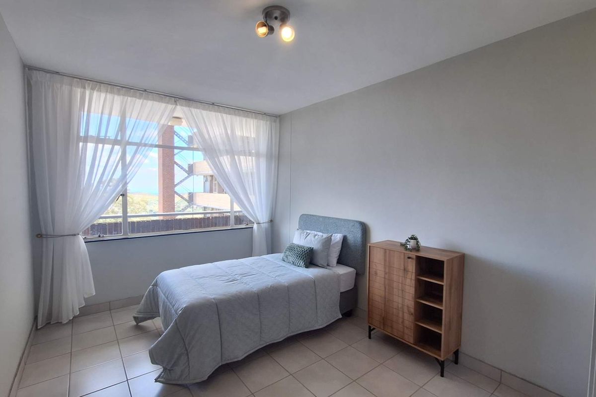 A bright bedroom featuring neutral tiled flooring, a large window allowing natural light, and a simple ceiling light fixture.