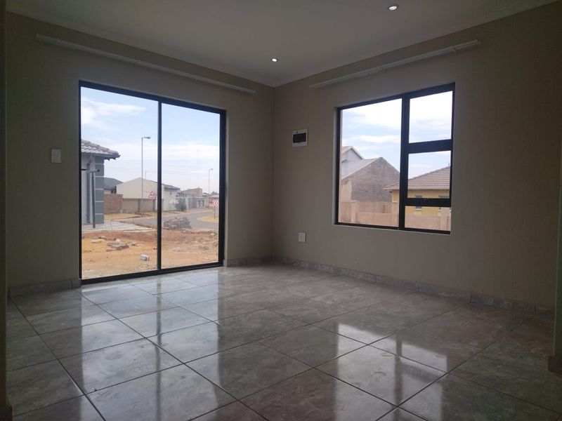 A bright, empty living area featuring modern tiled flooring, large aluminium-framed windows, and a sliding glass door providing natural light and outdoor access.