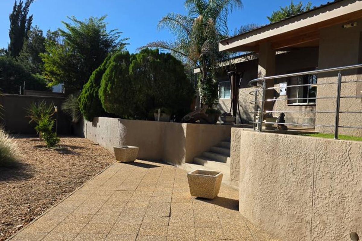 A well-maintained rear exterior area featuring a paved patio, retaining walls, and steps leading up to a covered veranda with stainless steel railings.