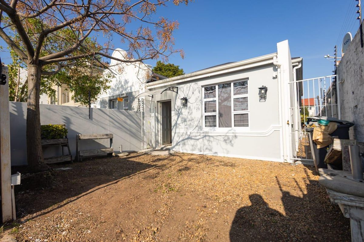 A rear exterior view of the property featuring a gravel courtyard, security fencing, and a simple facade with wall-mounted lighting.